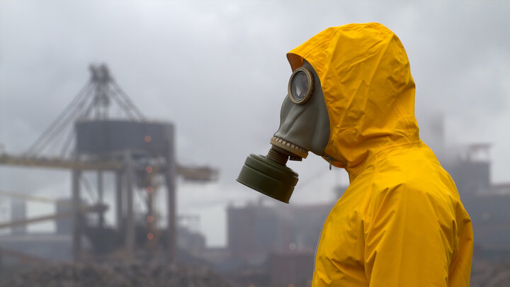 Zdjęcie przedstawia sylwetkę człowieka widzianą z profilu. Osoba ubrana jest w żółty skafander chroniący przed skażeniem. Twarz osoby zakrywa maska przeciwgazowa. W tle industrialny krajobraz.