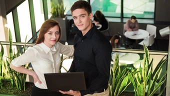 Na zdjęciu studentka i student stoją obok siebie na korytarzu uczelnianego budynku. Student trzyma w dłoniach laptop.