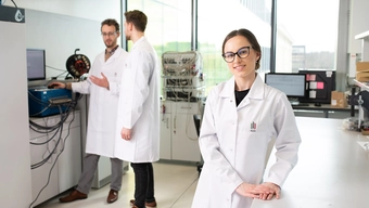 Trójka młodych naukowców w jasnym i nowoczesnym laboratorium. Na pierwszym planie kobieta w okularach uśmiecha się spoglądając w stronę widza.