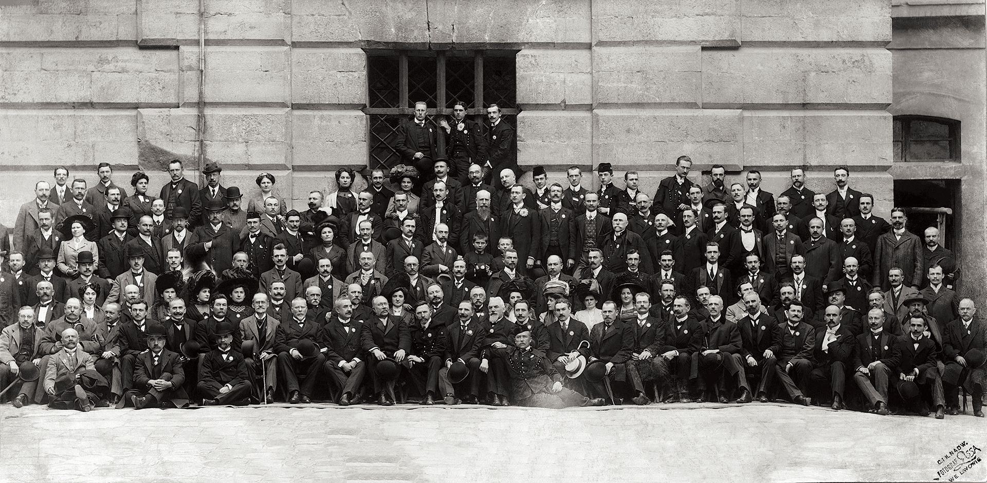 Archiwalne zdjęcie przedstawia bardzo liczną grupę ludzi siedzących oraz stojących w kilku rzędach przed ścianą budynku. Osoby pozują do zdjęcia. W przeważającej mierze grupę stanowią mężczyżni. Dostrzec można kilka kobiet.