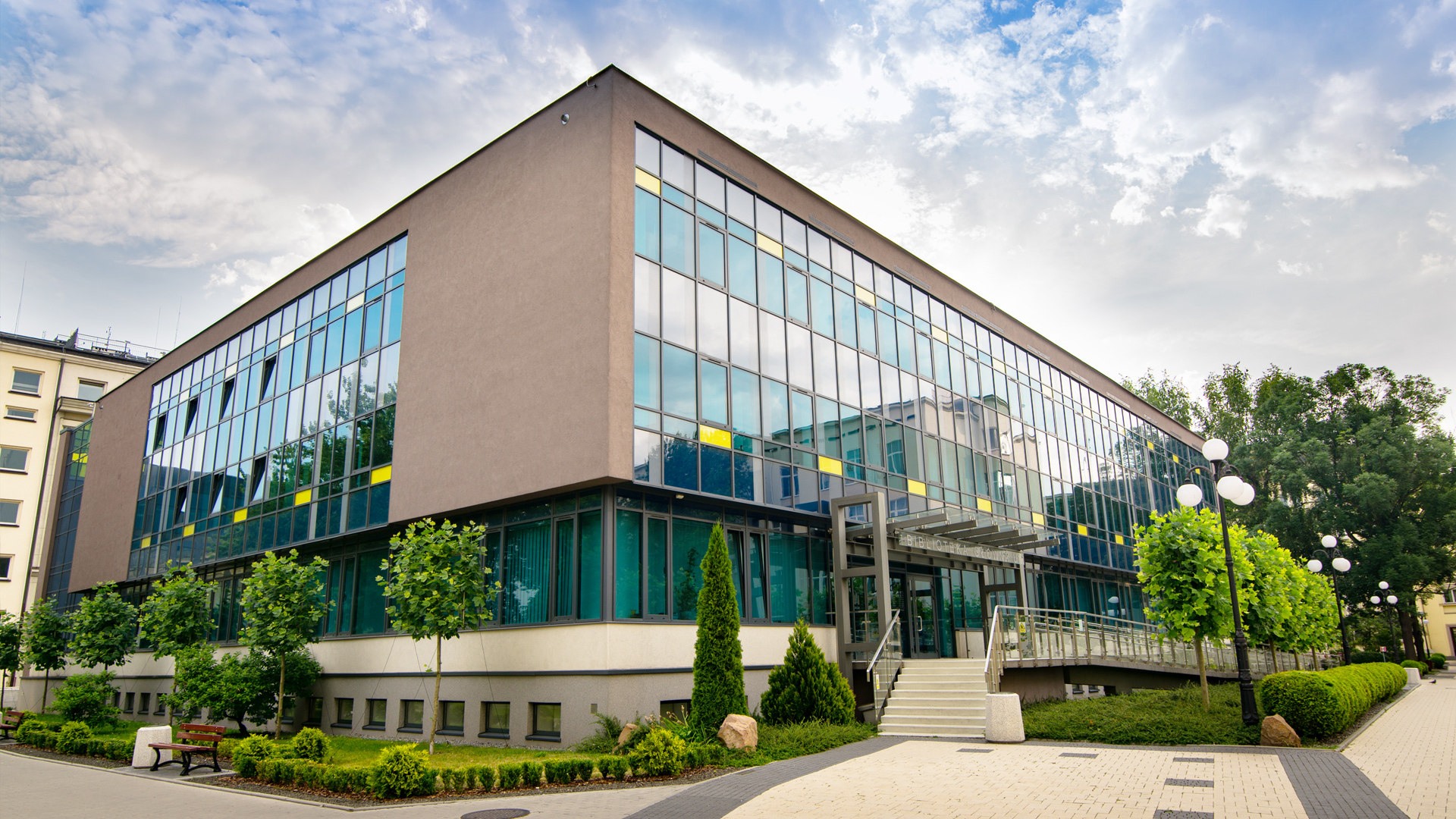 Zdjęcie dwukondygnacyjnego nowoczesnego budynku. W elewacjach dominują turkusowe odcienie okien z kontrastującymi żółtymi elementami. Elementy murowane są popielate. Dookoła budynku sporo zieleni.