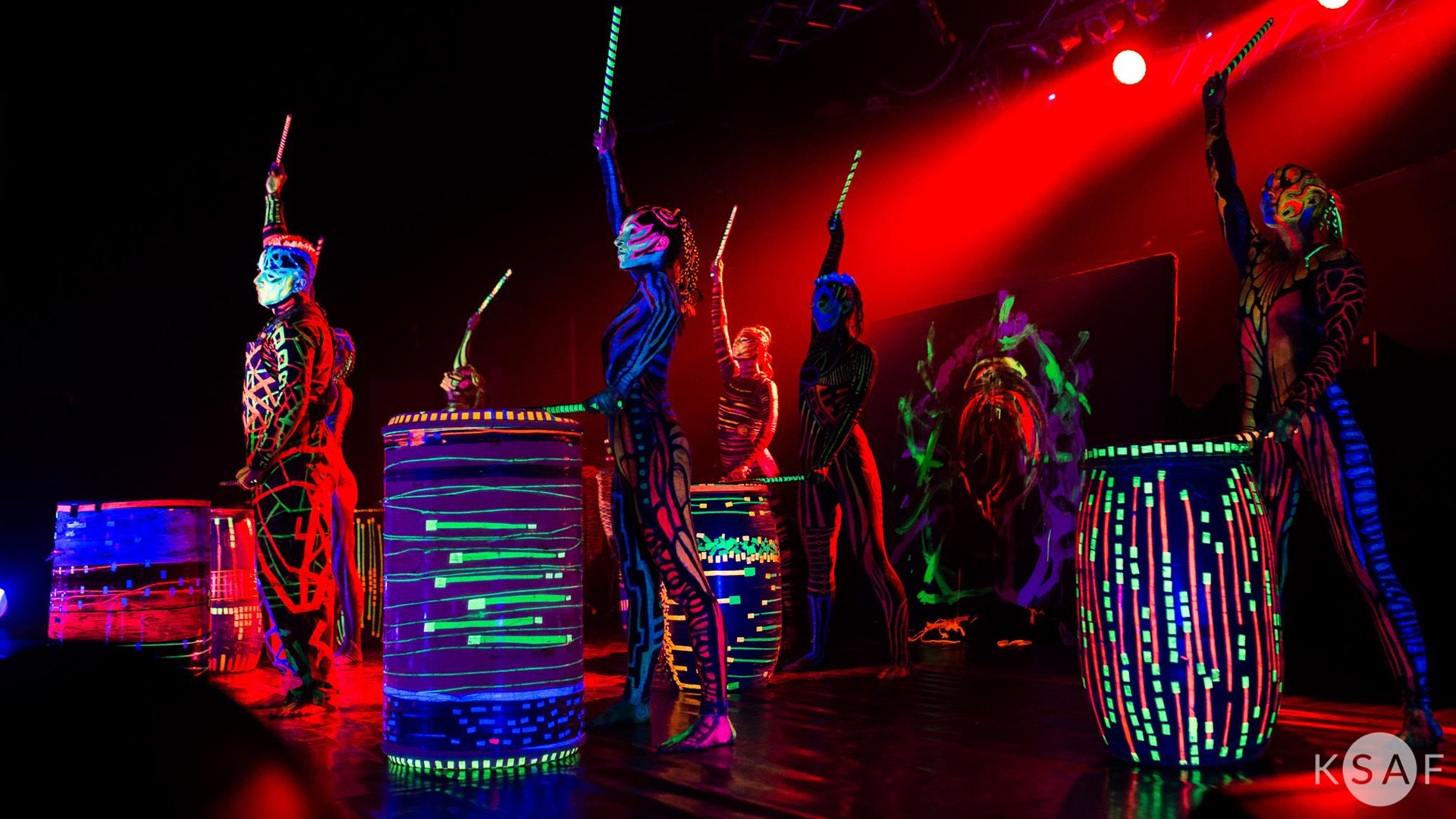 Zdjęcie występu scenicznego zespołu artystów stojących przez dużymi bębnami. Odziani są we wzorzyste kostiumy, których kolory widoczne są w świetle ultrafioletowym. Na scenie panuje półmrok kontrastujący z feerią barw kostiumów.