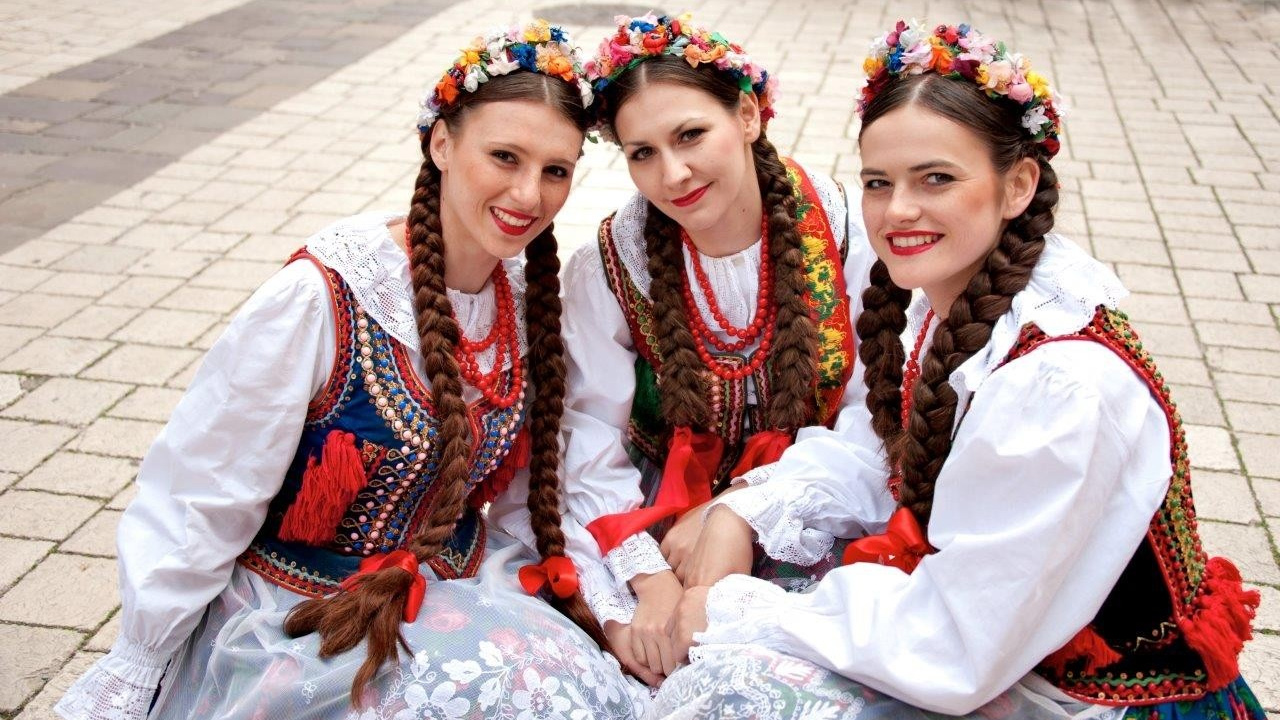 Zdjęcie trzech młodych kobiet w tradycyjnych strojach krakowskich. Ubrane są w białe koszule, granatowe i czarne kamizelki z kolorowymi haftami oraz czarne spódnice ozdobione kwiacistymi haftami. Na głowiach noszą kwieciste wianki, a na szyjach czerwone korale. Włosy zaplecione w warkocze, zwieńczone czerwonymi kokardami.