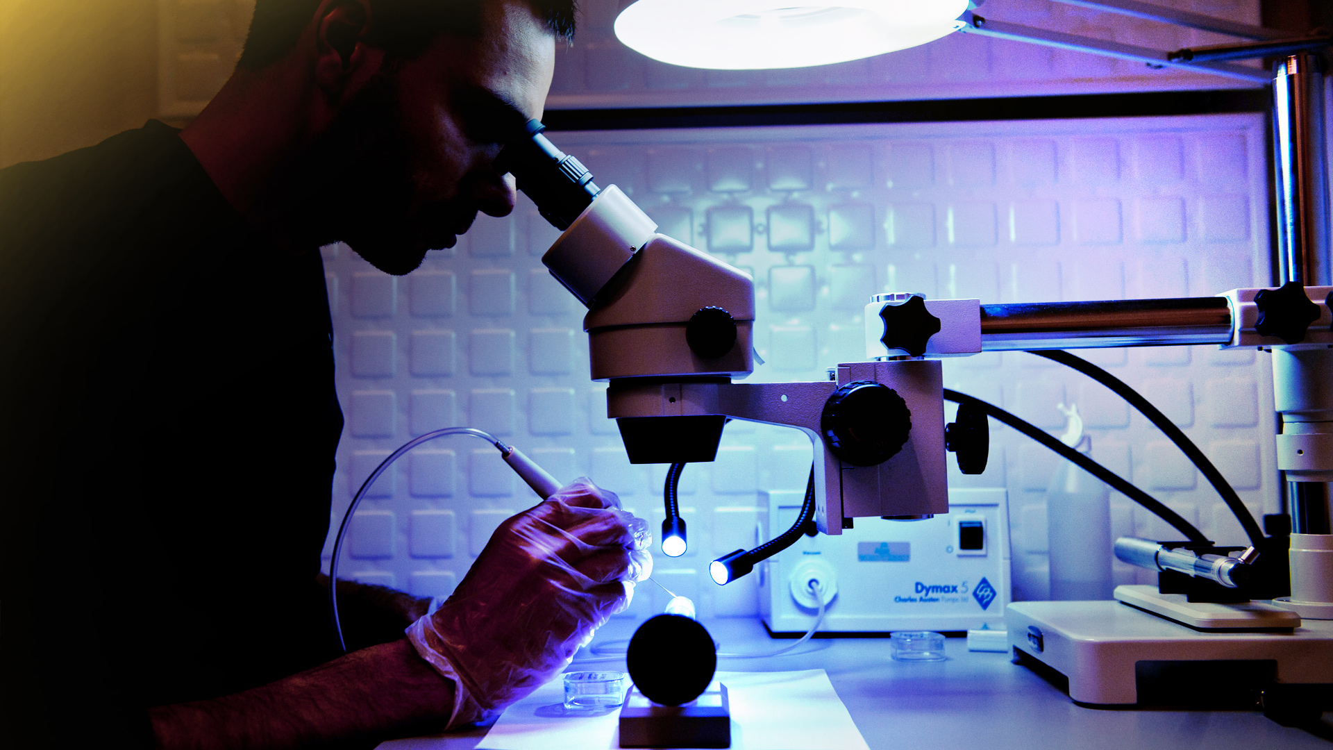 Mężczyzna widziany z profilu spoglądający przez mikroskop i analizujący próbkę. Próbka oświetlana przez dwie mocne lampki. 