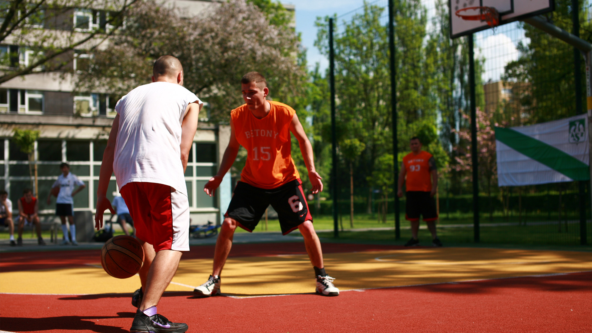 Zdjęcie dwójki koszykarzy grających w koszykówkę na tartanowym boisku, wykonane w ciepły słoneczny dzień. Powierzchnia boiska ma żółto-pomarańczowe kolory.