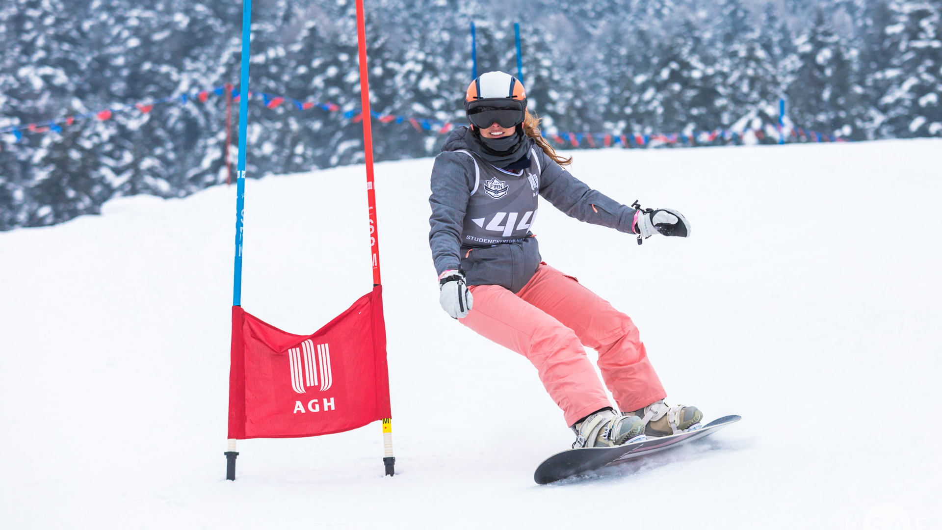 Zdjęcie zawodniczki jadącej w zakręcie na snowboardzie. W tle zimowa pogoda. Zawodniczka ubrana jest w szarą kurtkę oraz czerwone spodnie. Na głowie nosi jasny kask oraz ciemne gogle. Mija podwójną tyczkę z nadrukowanym na czerwonym materiale logotypem AGH.