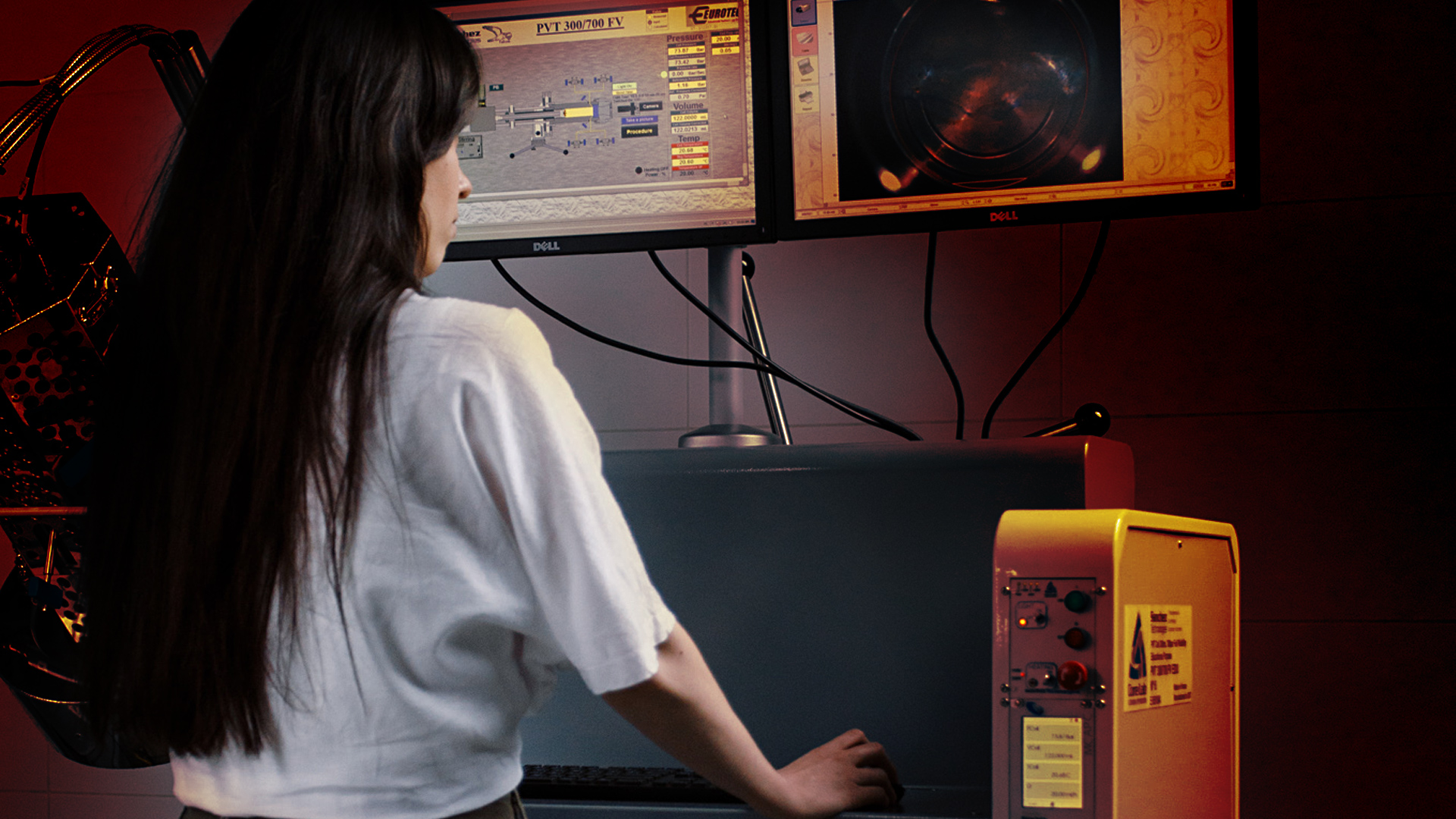 Zdjęcie kobiety stojącej przed aparaturą badawczą. Widzimy jej plecy. Ma długie ciemne włosy i jasny t-shirt. Operuje myszką. Przed nią na wysokości jej oczu wiszą dwa monitory. 
