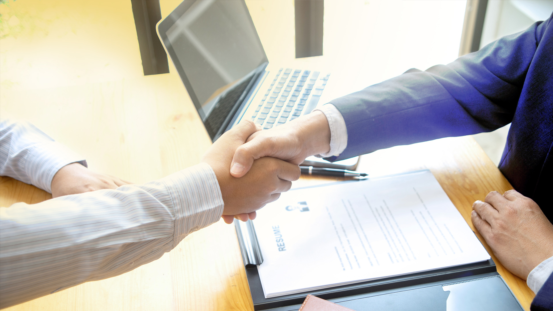 A close-up on a handshake of two people. On the desk below, there is a laptop and a file with documents.