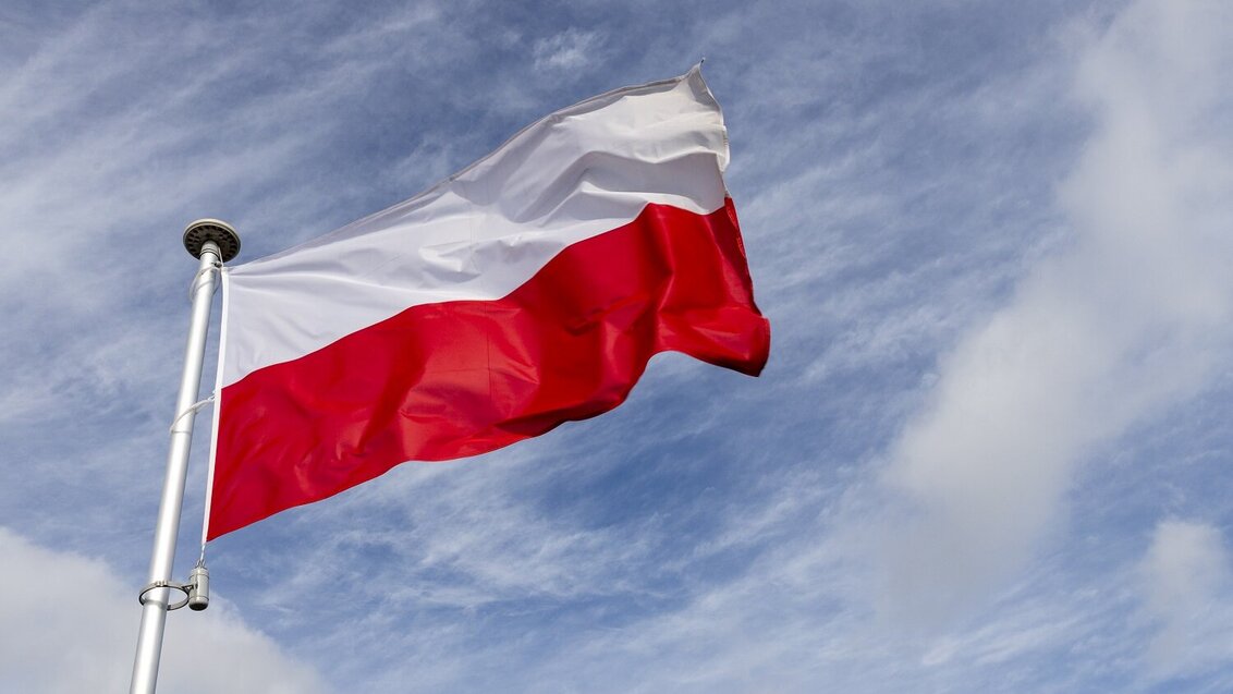 Na zdjęciu biało-czerwona flaga Polski, powiewa na wietrze, na tle nieba.