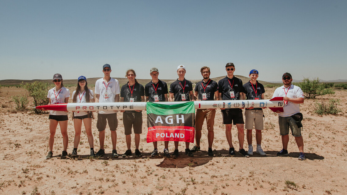 Na zdjęciu dziesięcioro członków zespołu AGH Space Systems. Stoją obok siebie w rzędzie, trzymając przed sobą rakietę. Na rakiecie zawieszona flaga AGH.