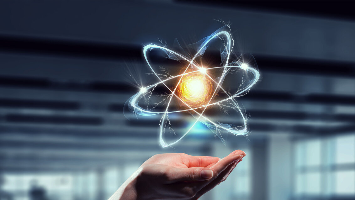 Abstarakcyjna grafika przedstawiająca damskie dłonie nad którymi unosi się atomow z widocznymi protonami w rozgrzanym jądrze oraz błękitnymi orbitami po których krążą elektrony.