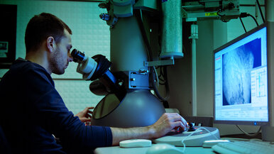 Młody mężczyzna spoglądający w okular mikroskopu. Po prawje stronie widać monitor komputerowy.